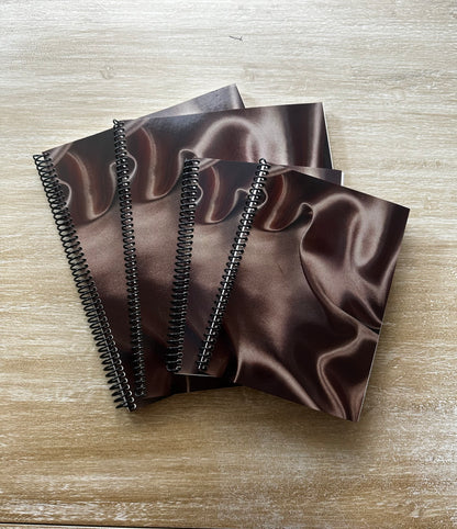 Four spiral-bound notebooks with a dark brown satin-patterned cover, arranged in a fan shape on a light wood surface.