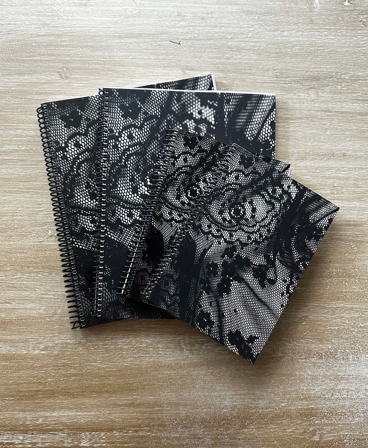 Four black spiral-bound notebooks with black lace-patterned covers are stacked on a light wood surface.