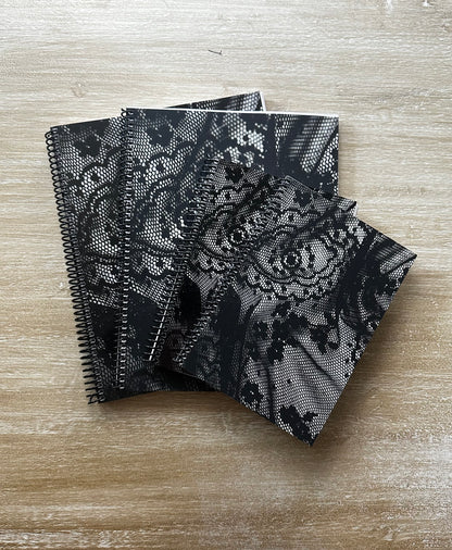 Four black spiral-bound notebooks with black lace-patterned covers are stacked on a light wood surface.